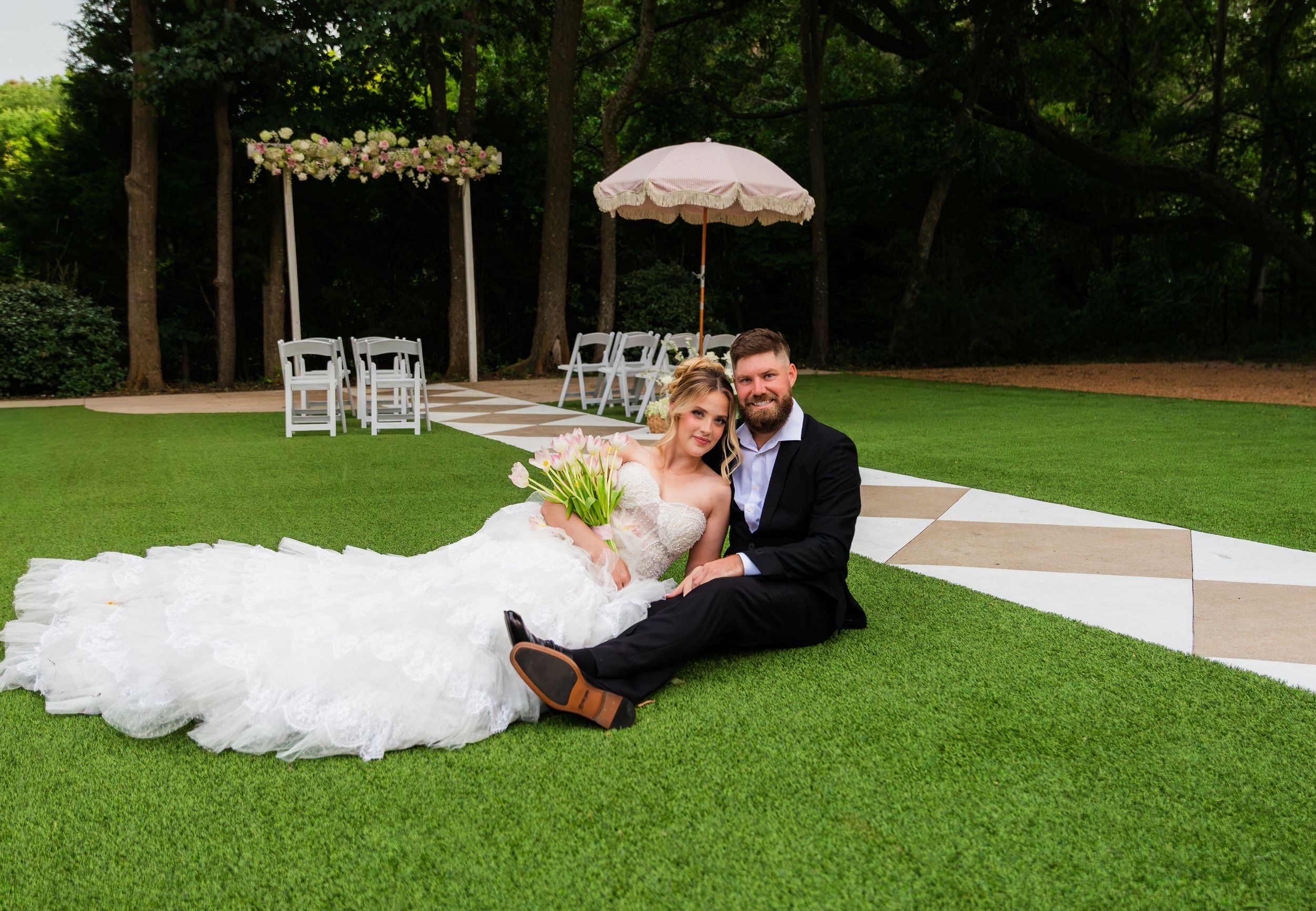 A bride and groom sit together on lush green grass at an outdoor wedding venue with a checkered dance floor and floral arch.