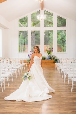 Bride in strapless ballgown with bouquet stands in elegant white chapel with large windows and rows of chairs.