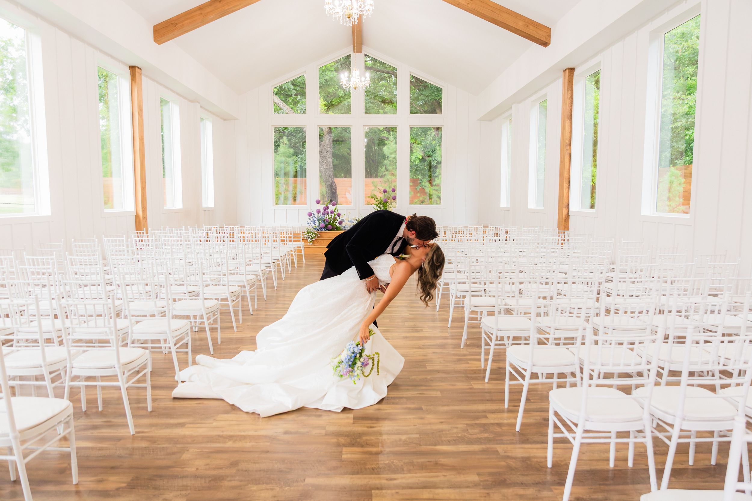 Bride and groom share a romantic dip kiss in a bright white wedding chapel with rows of white chairs.