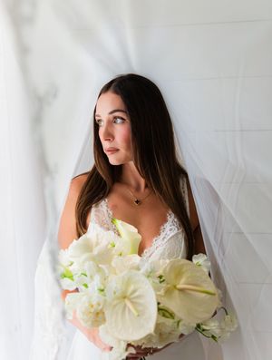 Bride in lace wedding gown holding white floral bouquet with veil, gazing away in elegant bridal portrait.
