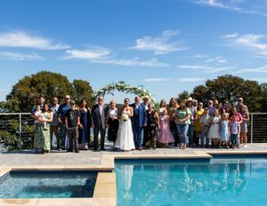 Wedding party and guests gather for a group photo by a pool with a floral arch backdrop. Eagle Mountain Fort Worth Texas Wedding.