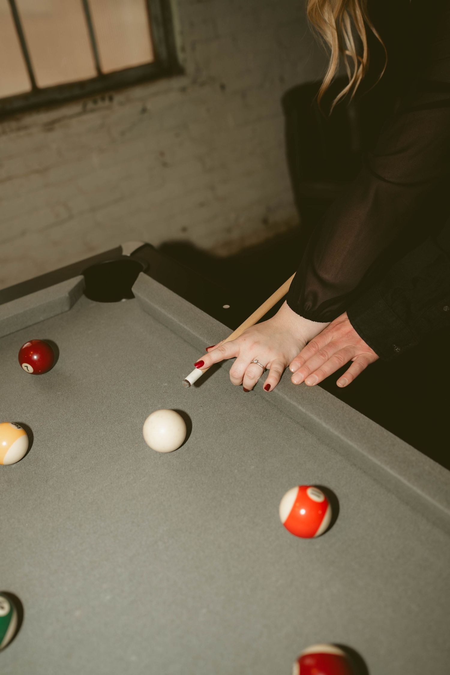 Hands holding a pool cue over a gray billiards table with scattered colorful balls in dim lighting during an engagement session in Dayton OH