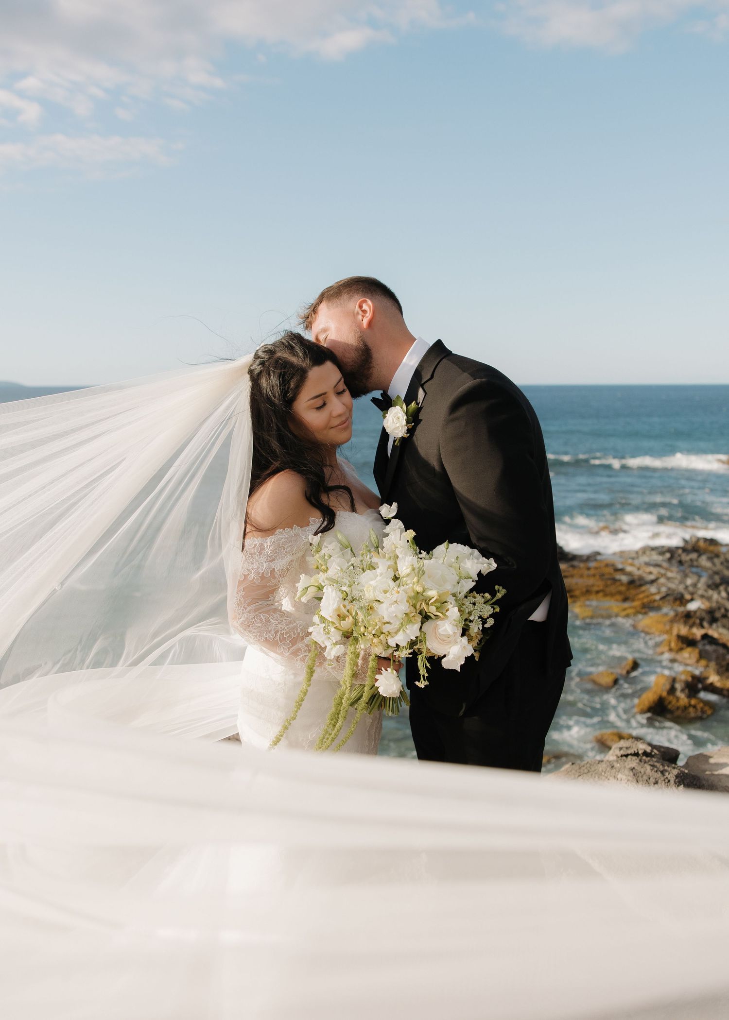 A romantic wedding couple shares a tender moment by the ocean as the bride's veil flows in the breeze.