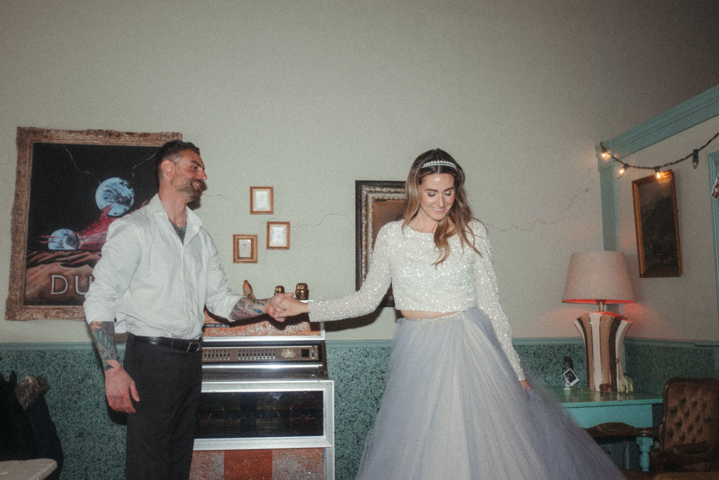 A couple dances together in a vintage-style room with a teal wall and pink lamp.