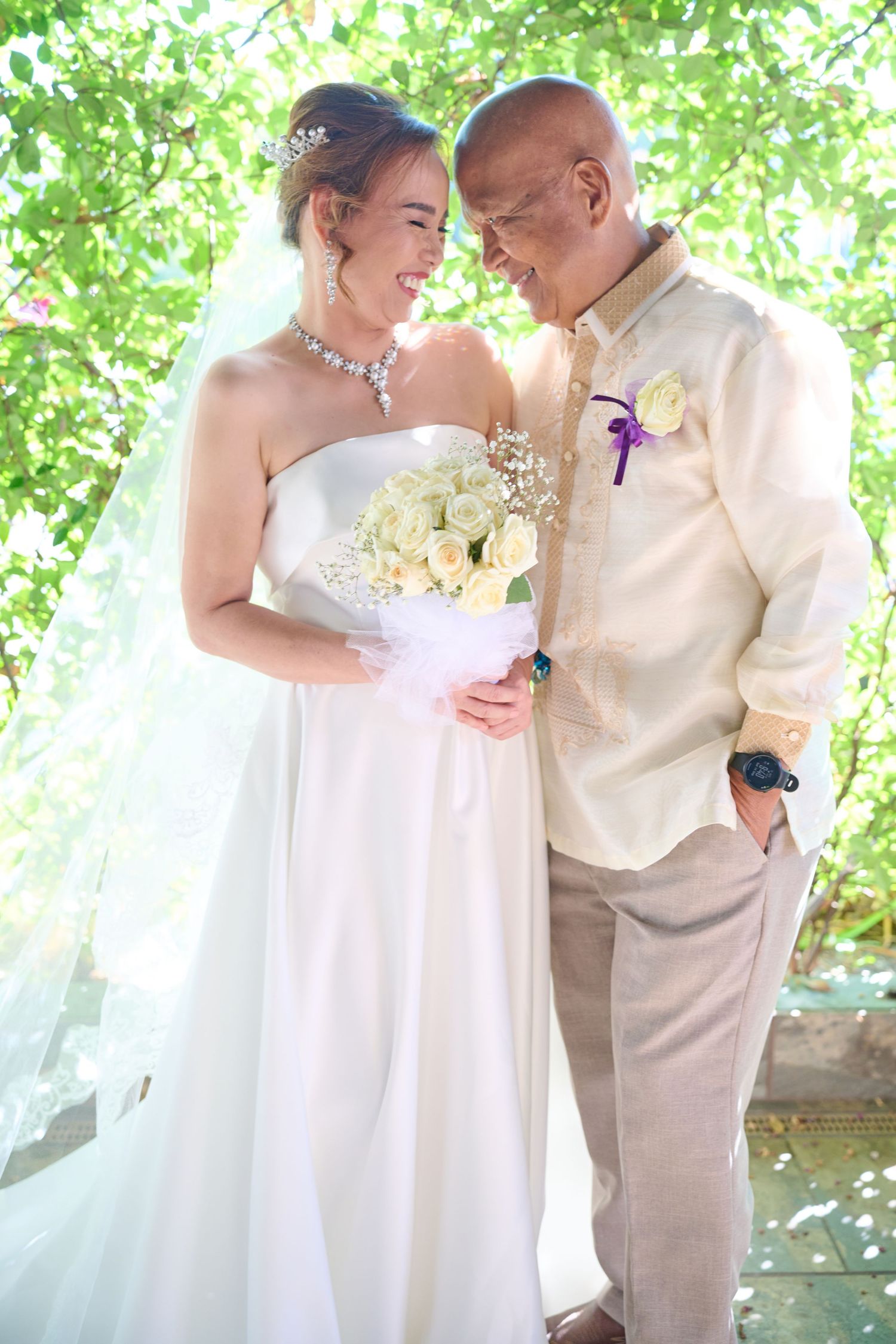 A joyful wedding moment outdoors with a white bridal gown and bouquet of white roses under sunlit green foliage.