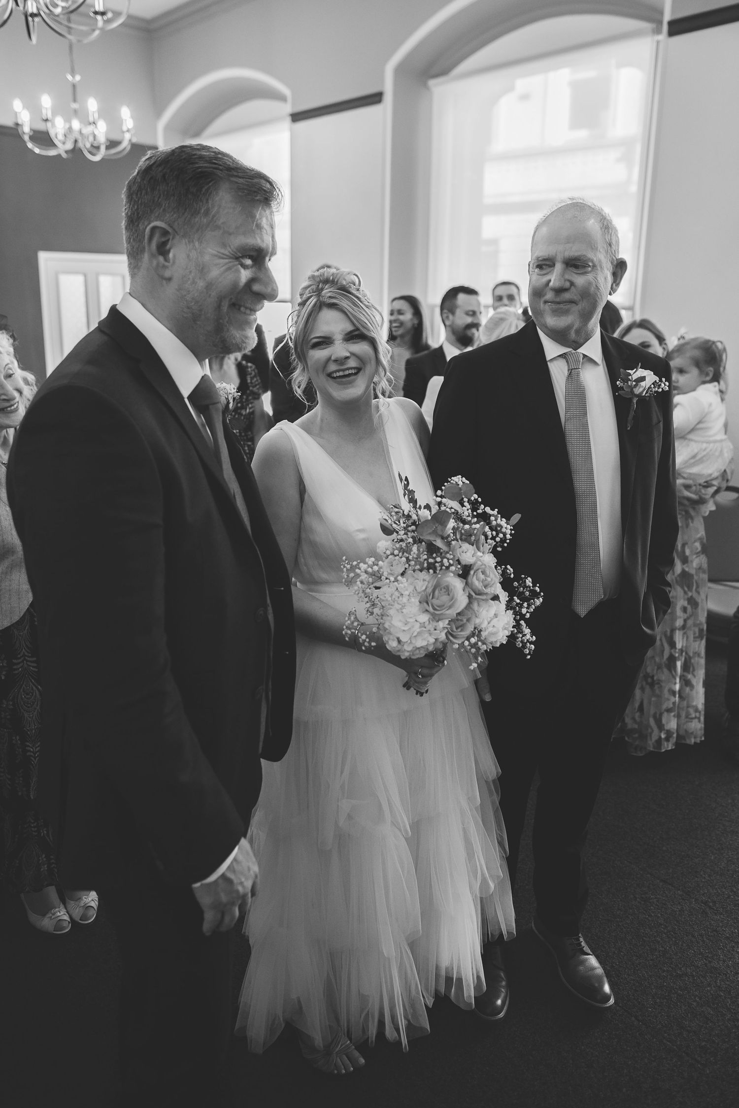 A black and white wedding photo showing a bride in a flowing tulle dress with her bouquet and two men in suits.
