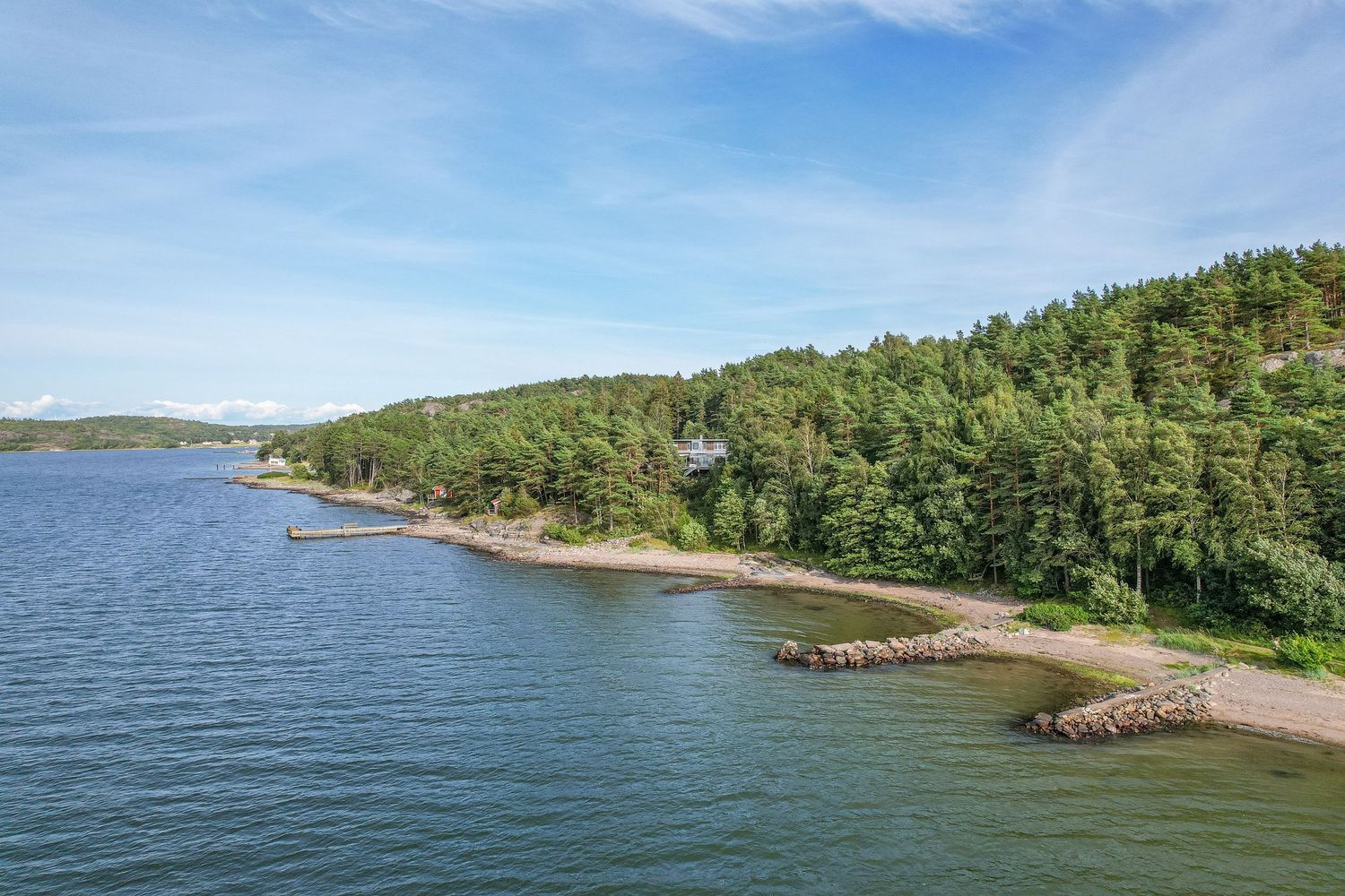 Scenic coastal view of forested shoreline with rocky outcrops along blue water under summer sky.