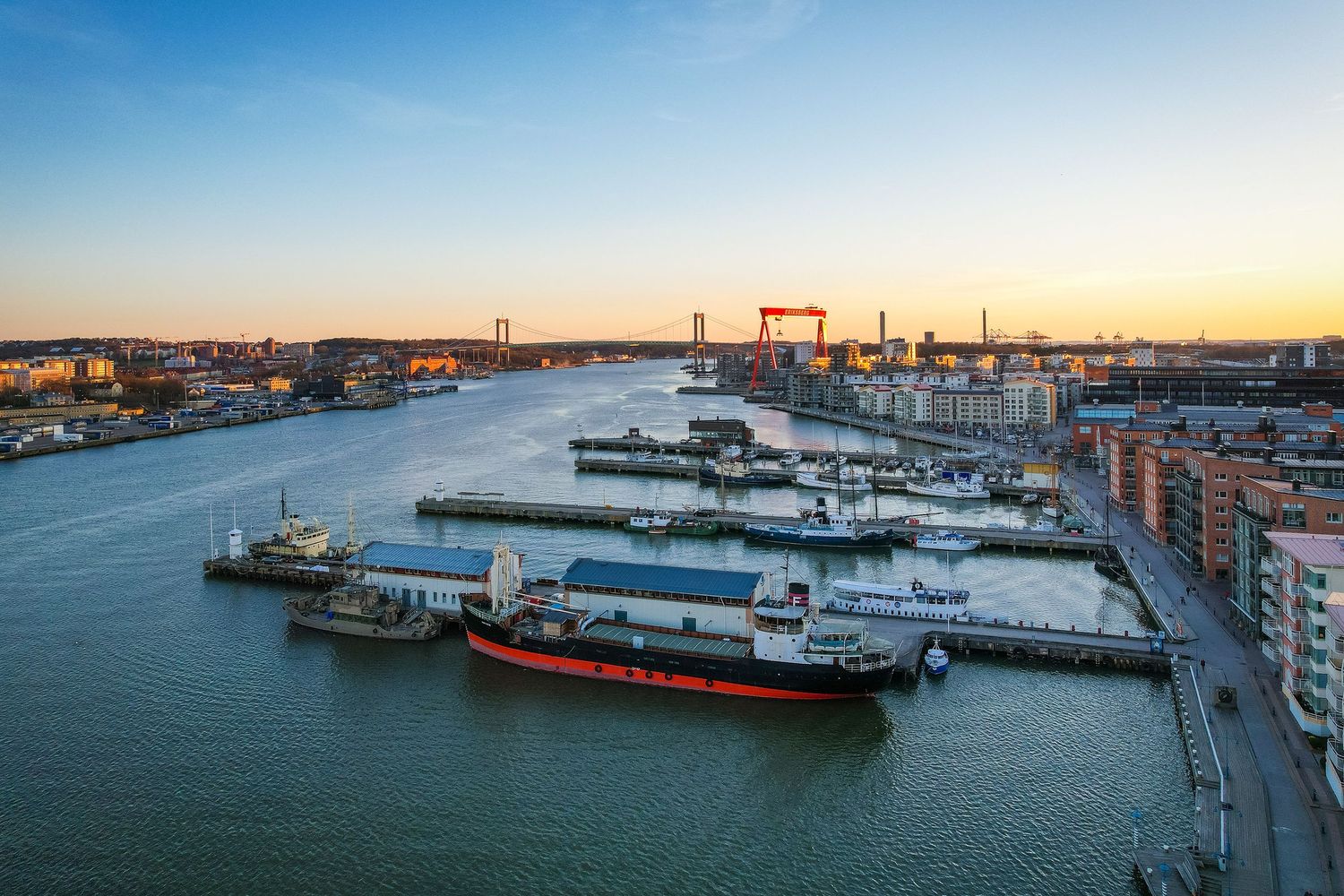 Ships docked at Gothenburg harbor with industrial cranes and city buildings along the waterfront during golden hour.