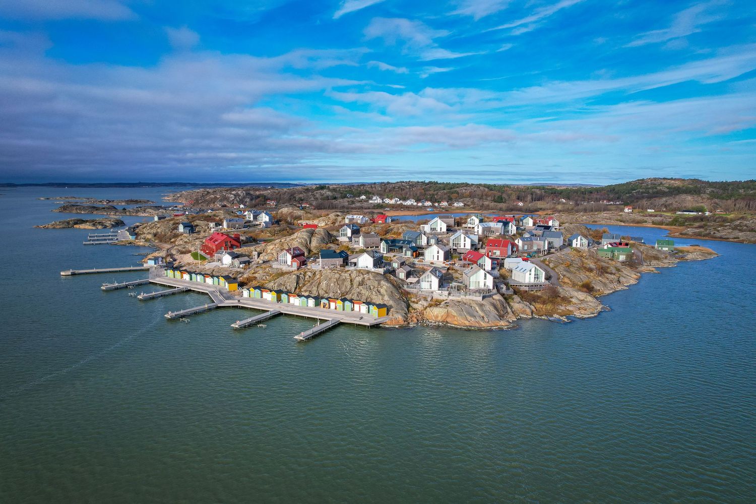 Aerial view of a small coastal fishing village with colorful houses and boats docked along rocky shoreline under blue skies.