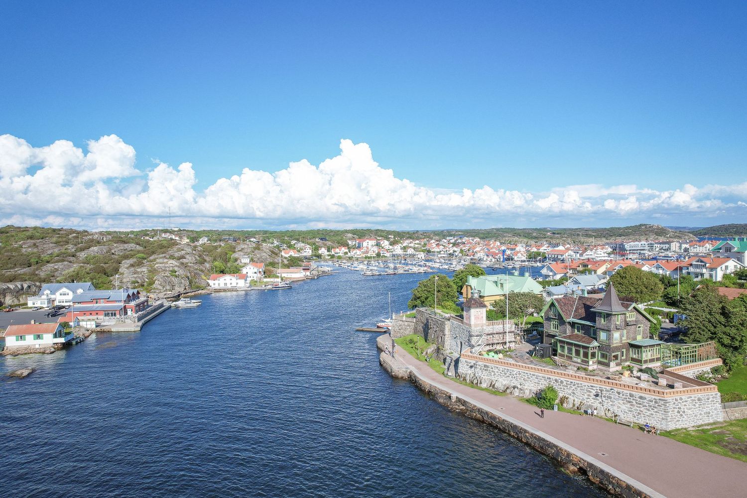 Aerial view of a scenic waterfront town with boats docked along a curved harbor on a sunny summer day.