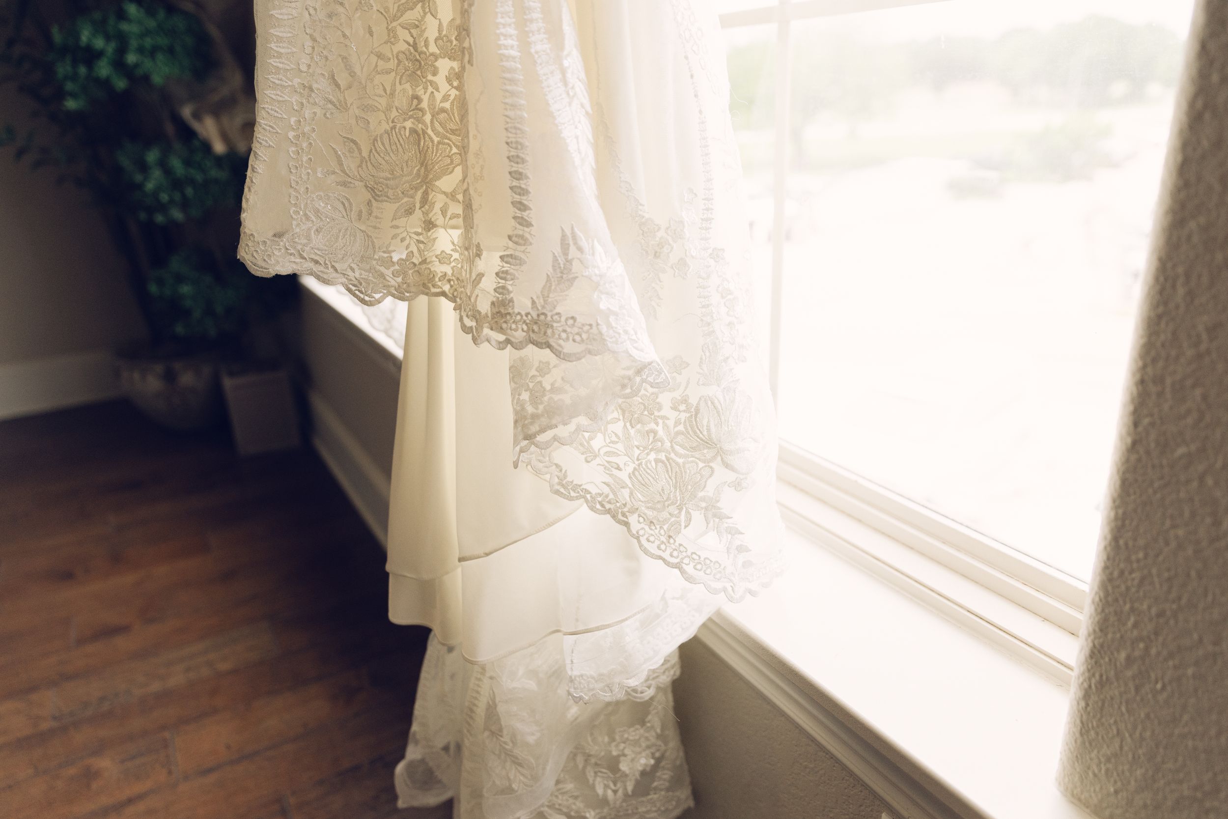 Sunlit white lace wedding dress hanging by window.