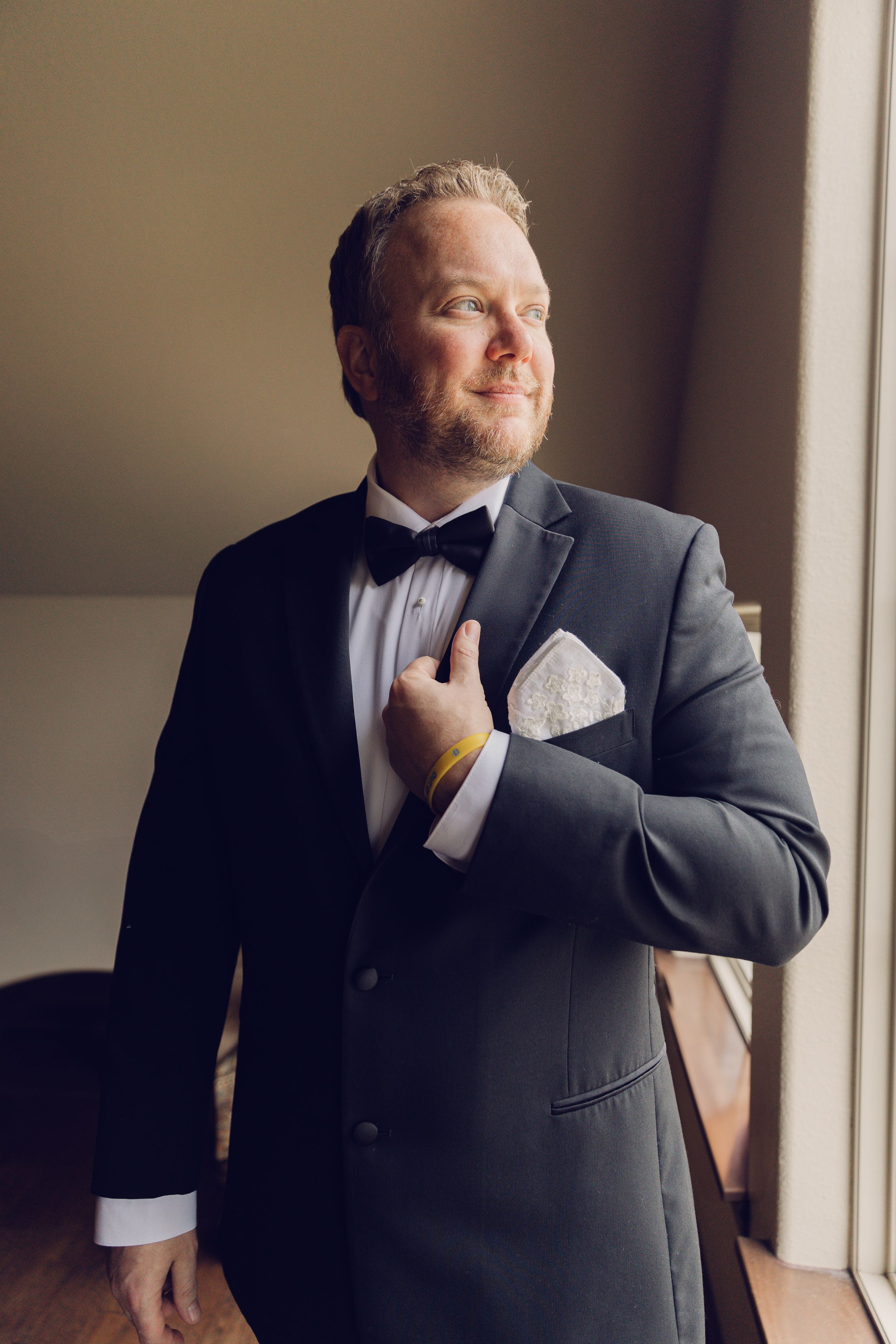 Person in formal black suit adjusting tie near window.