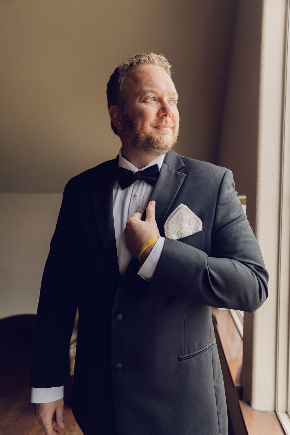 Person in formal black suit adjusting tie near window.