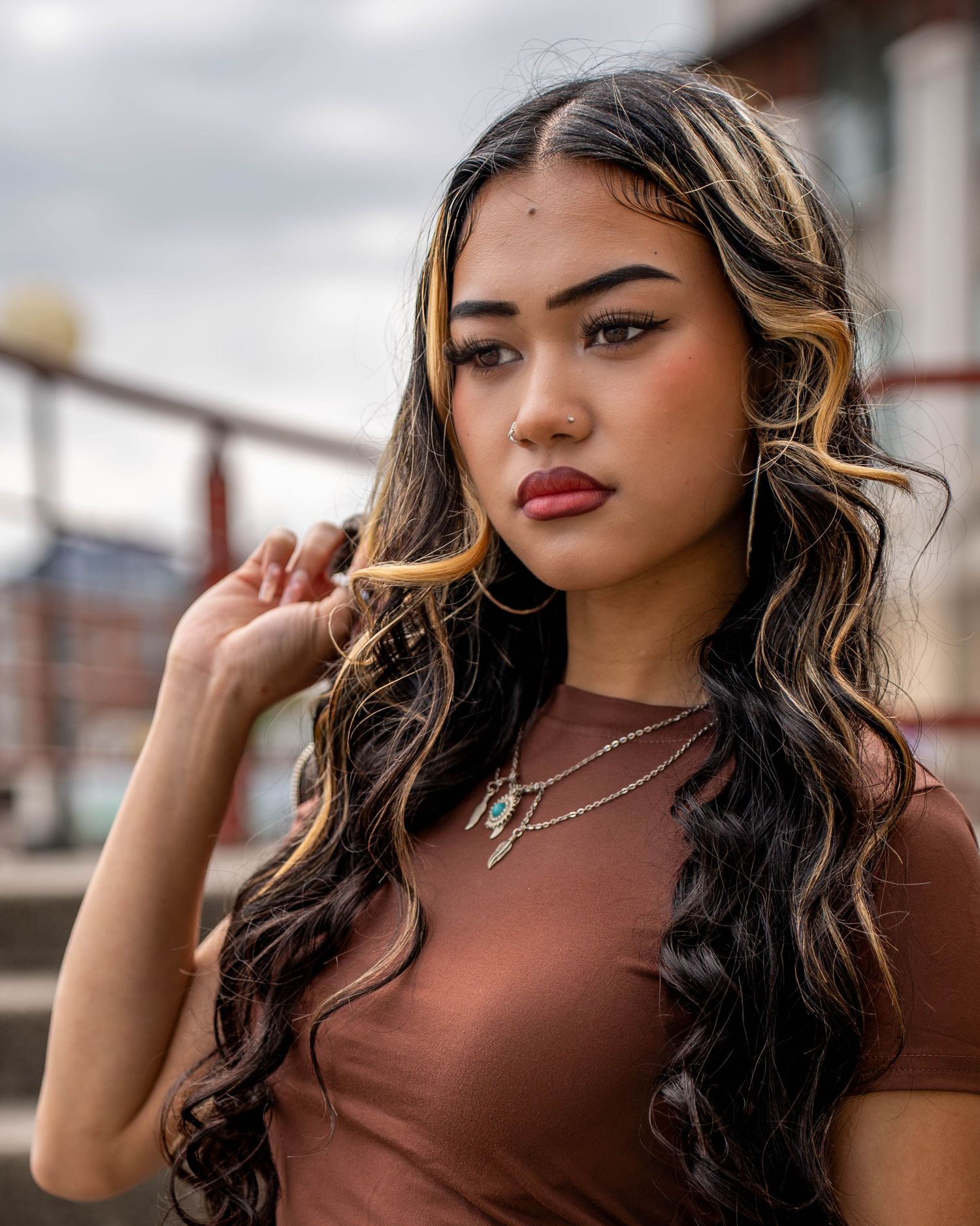 Portrait of model in brown top wearing necklace against cloudy sky.