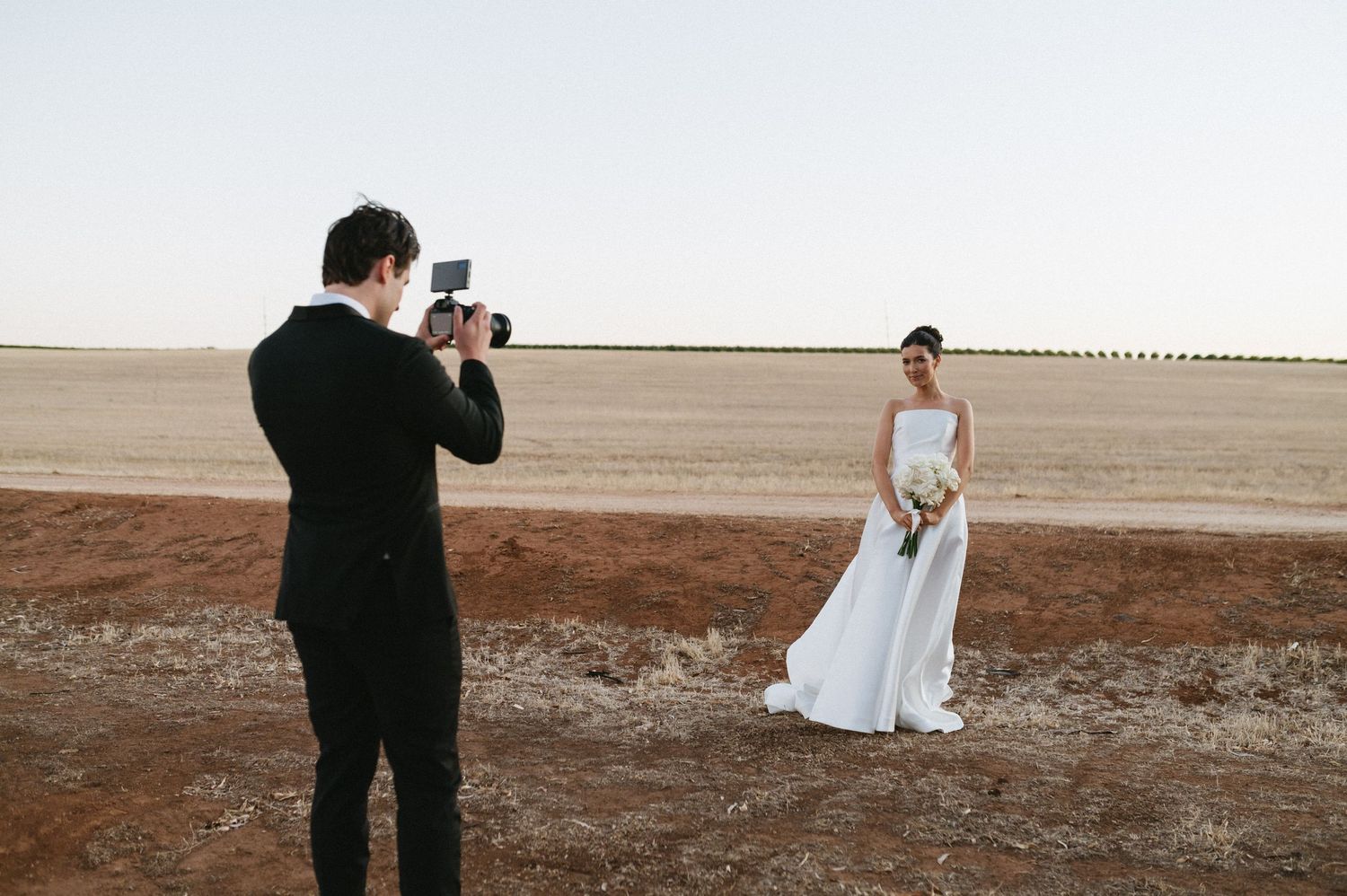 A photographer captures a bride in white dress standing in a vast open field.