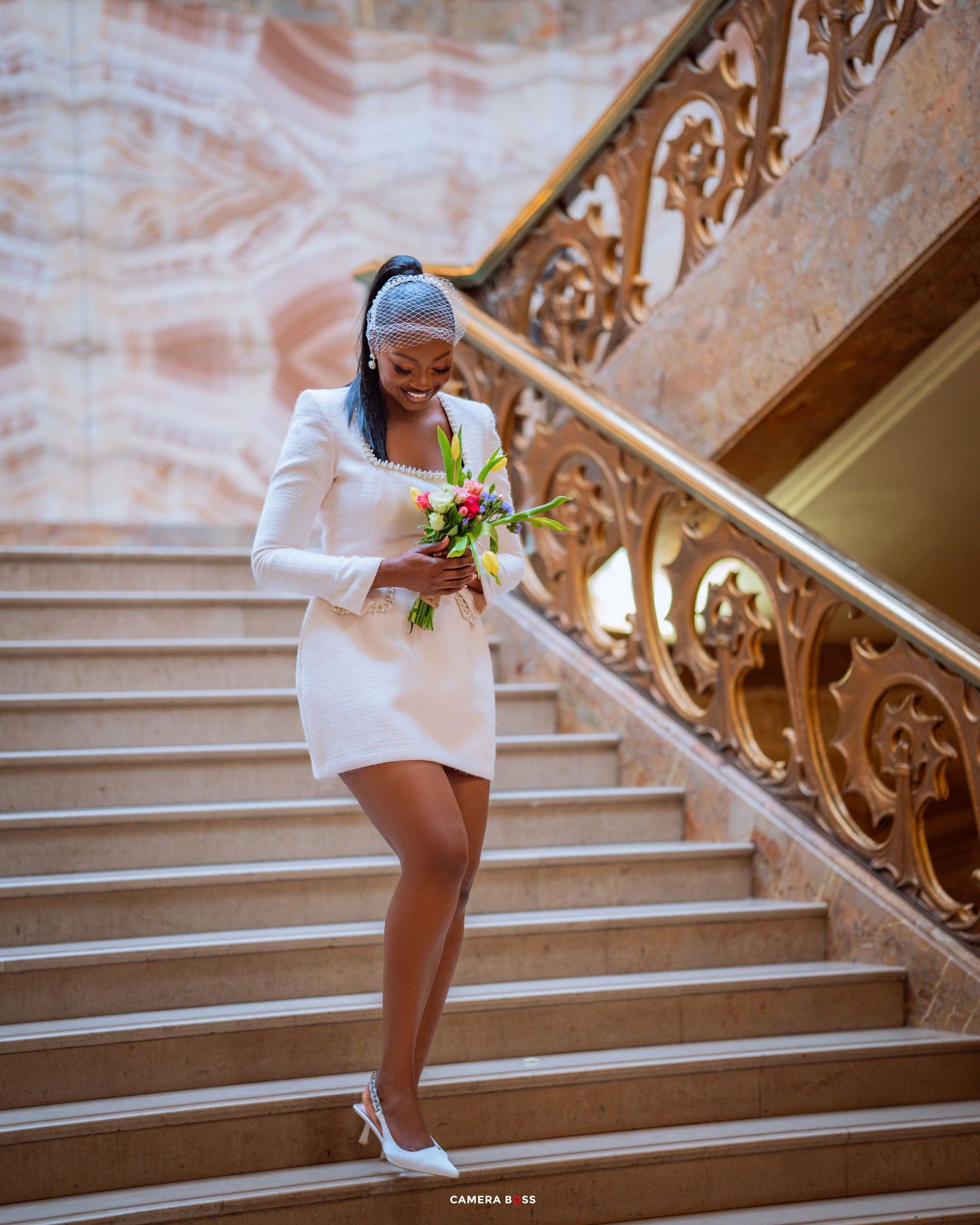 A person in a white dress and sneakers poses elegantly on an ornate grand staircase while holding a small bouquet of flowers.