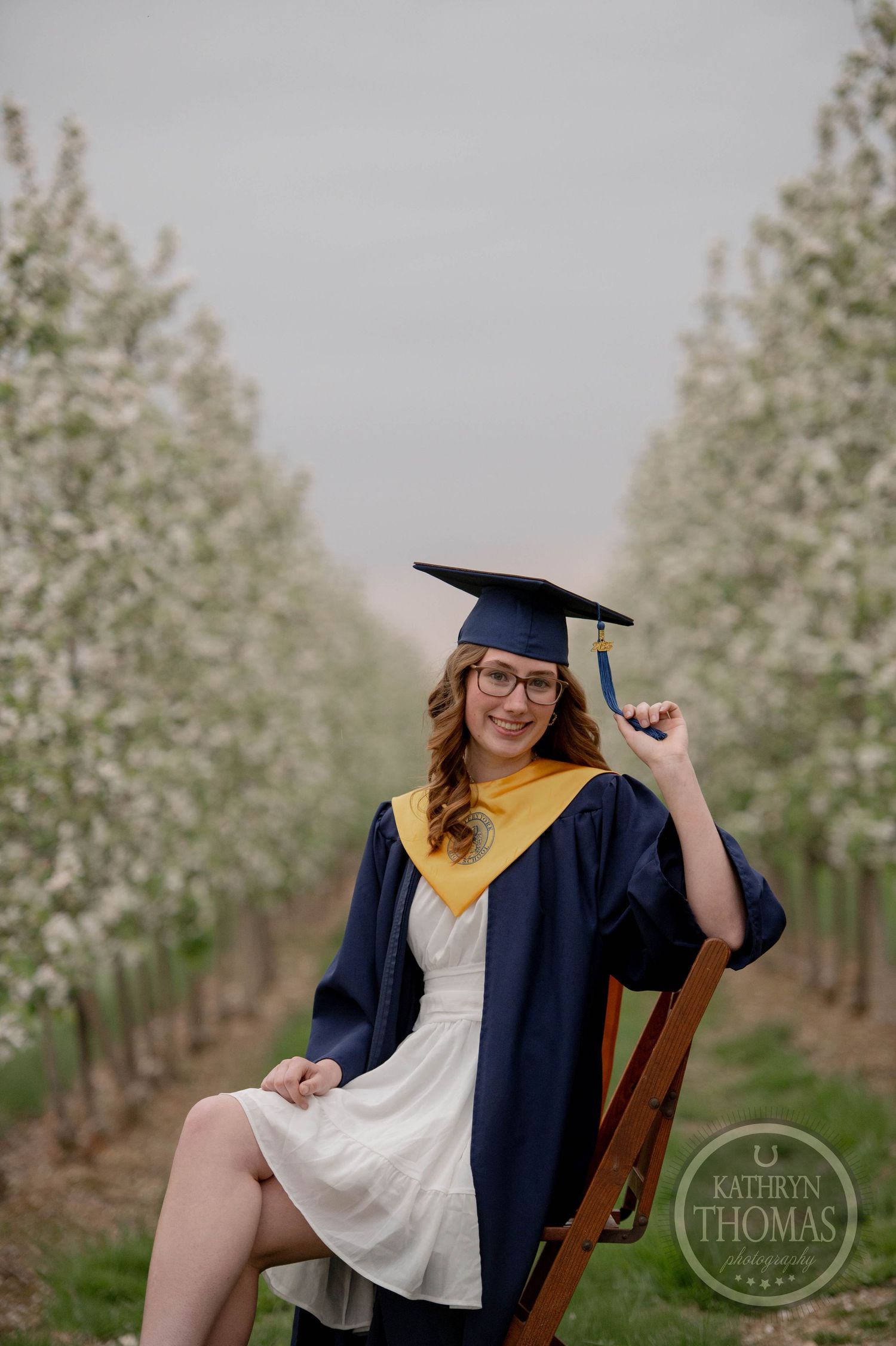 Graduate in cap and gown posing thoughtfully in blooming spring orchard during outdoor photo session.