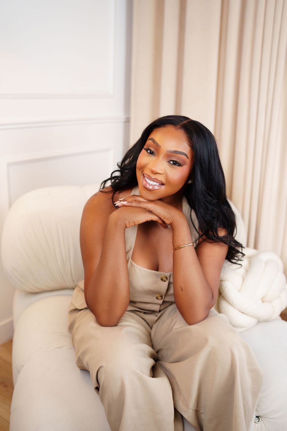 A person smiles while posing on a cream-colored couch in a white room with soft natural lighting.