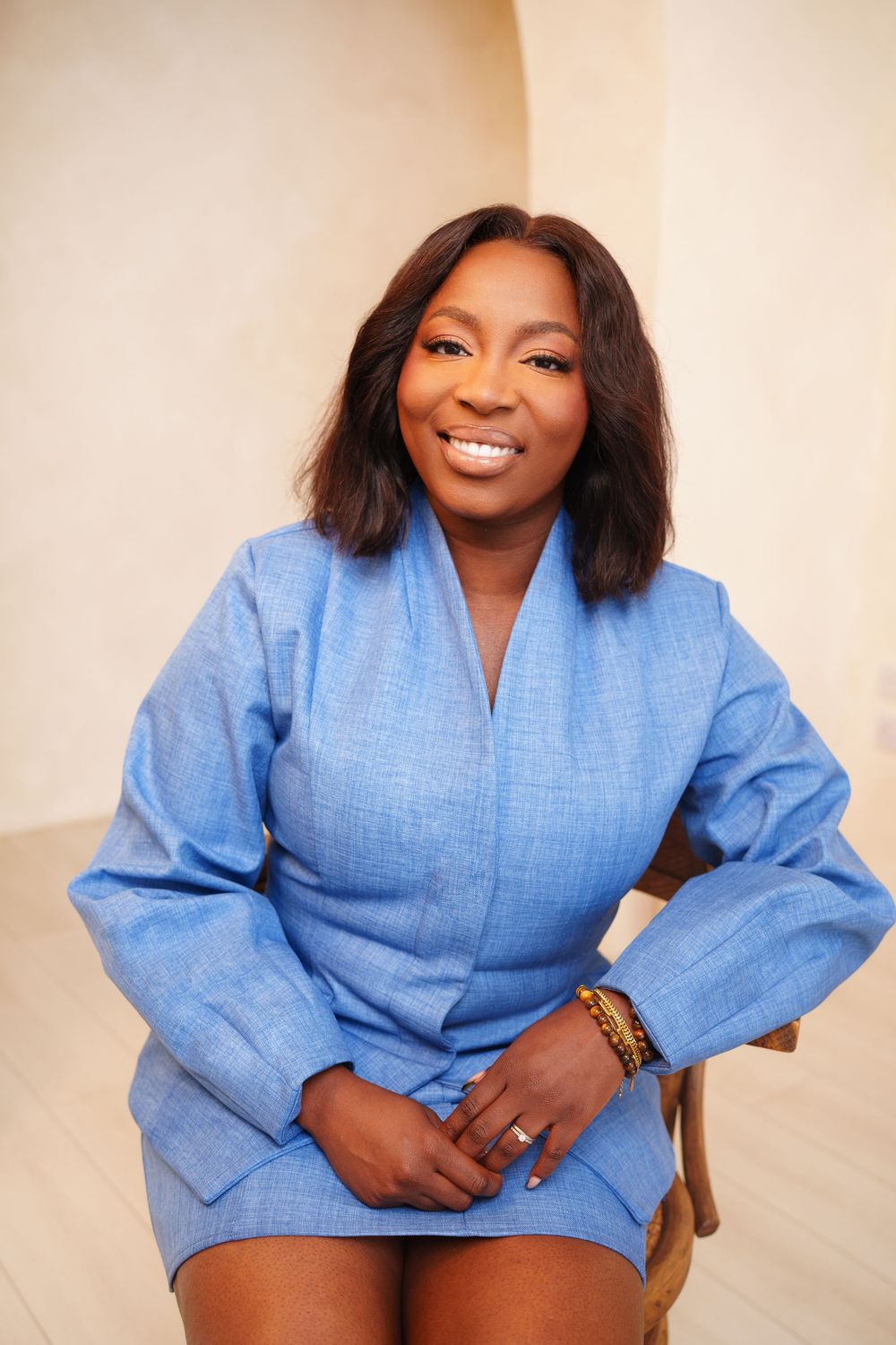 Person wearing a light blue wrap-style jacket dress and bracelet poses against a beige background with a warm smile.