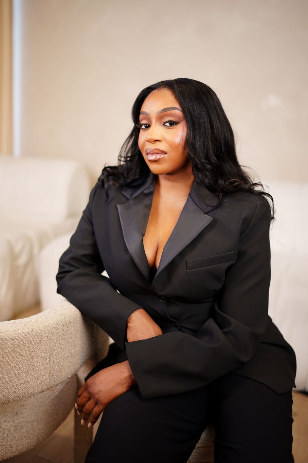 A portrait of someone in a black blazer sitting on a beige couch with soft lighting and elegant makeup.
