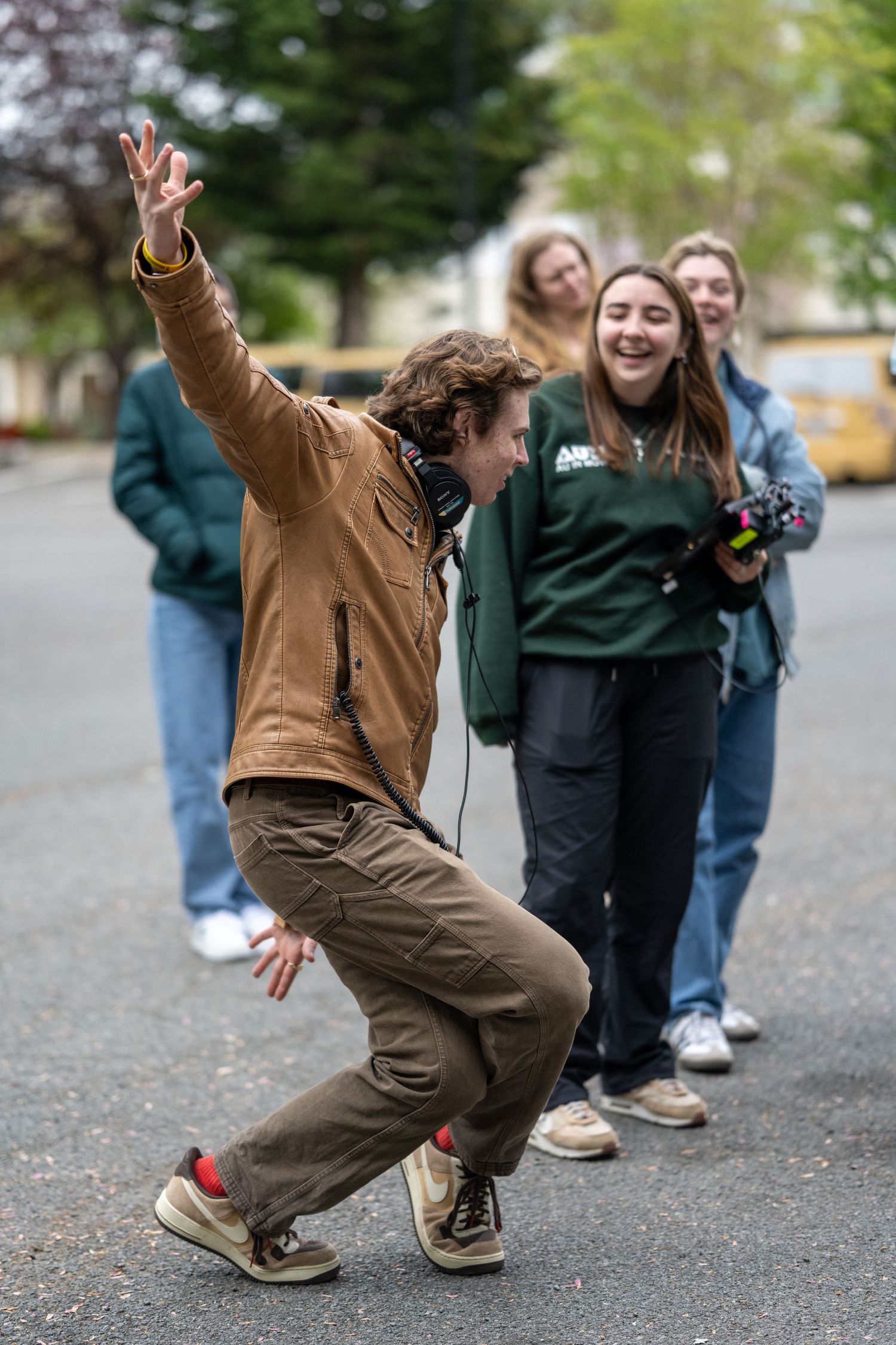 Person in brown jacket dancing energetically on street while others watch and laugh.