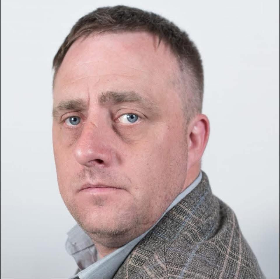 A middle-aged man in a grey tweed jacket looks to the side with a serious expression against a white background.