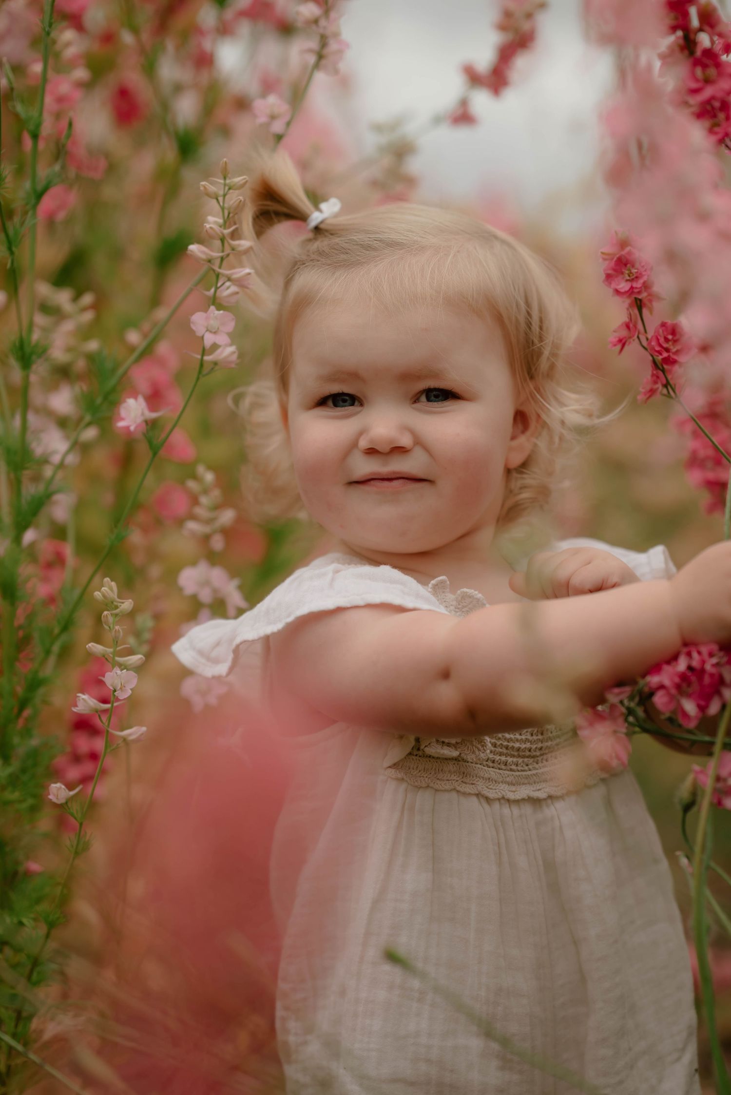 A small child in a white dress stands joyfully among pink and white blooming flowers in a dreamy garden setting.