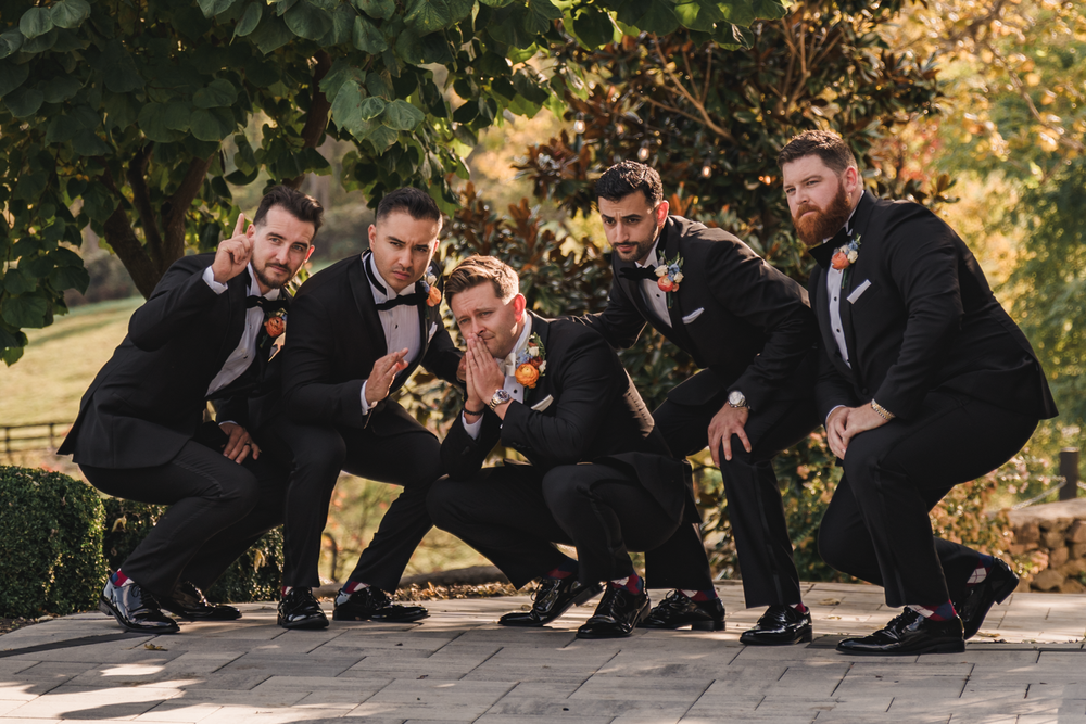 Groomsmen in black suits crouching and laughing together outdoors during autumn wedding photoshoot.