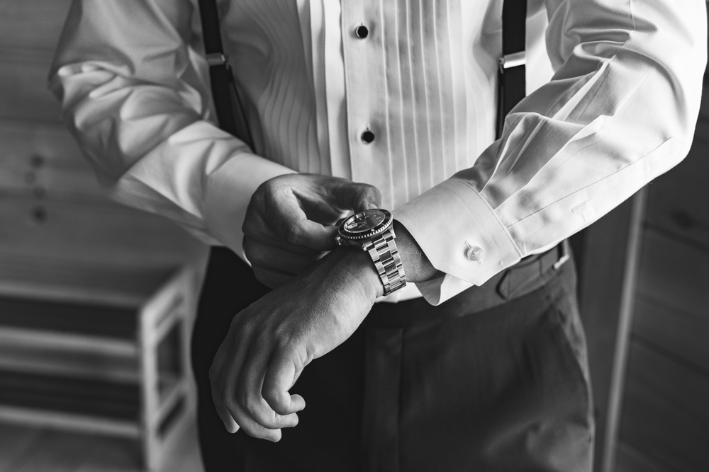 A person in formal attire checks their wristwatch while wearing a tuxedo shirt and dark trousers.