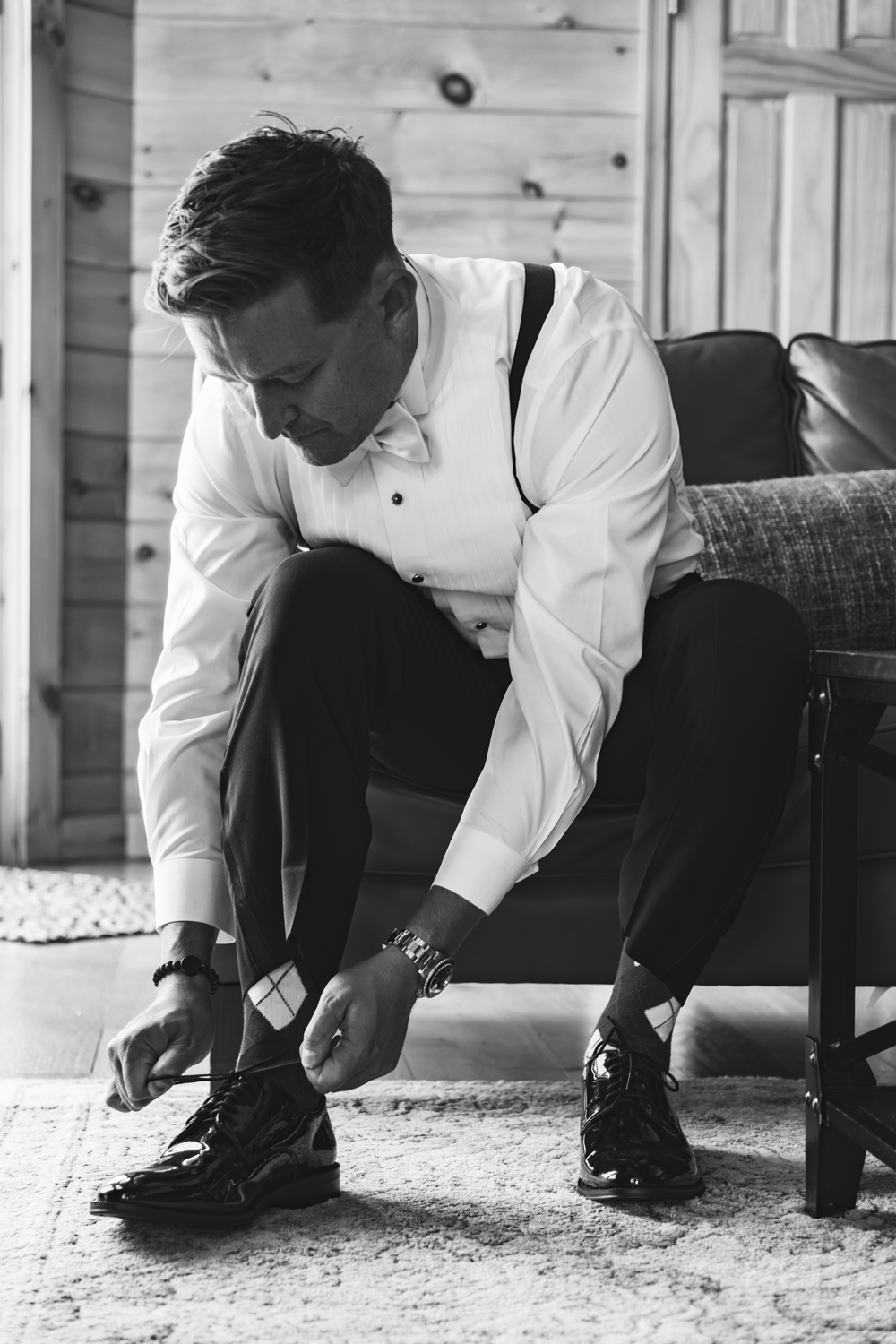 Black and white photo of someone in formal attire sitting on a couch while tying dress shoes.