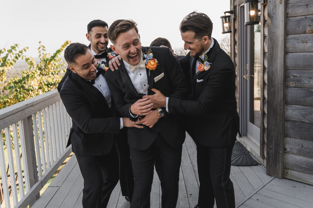 Four groomsmen in black suits sharing a lighthearted moment on a porch balcony.