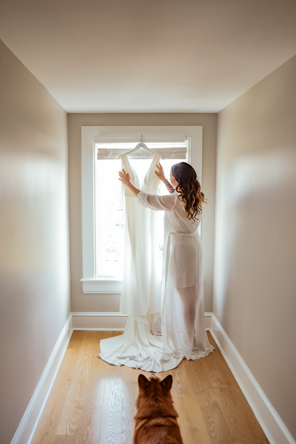 A person in a white robe adjusts a wedding dress hanging in a window while a dog watches from the hallway.