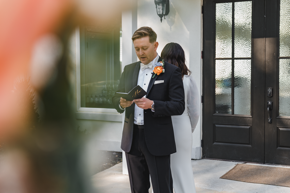 A couple stands close together on a porch reading from papers during what appears to be a wedding ceremony.
