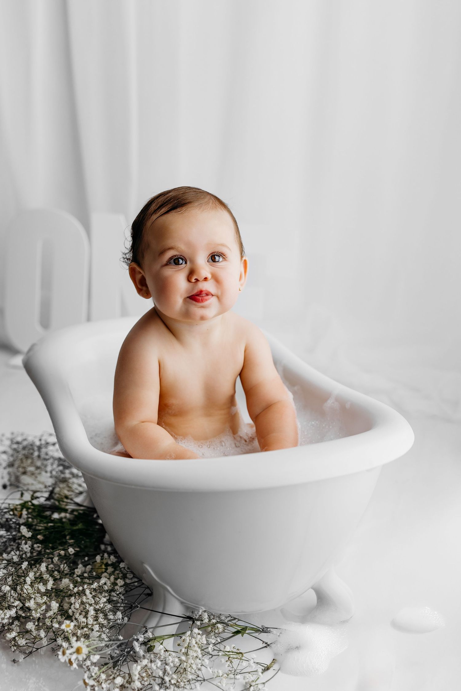Baby wearing floral crown plays with maracas while sitting in white bathtub during bright indoor photoshoot.
