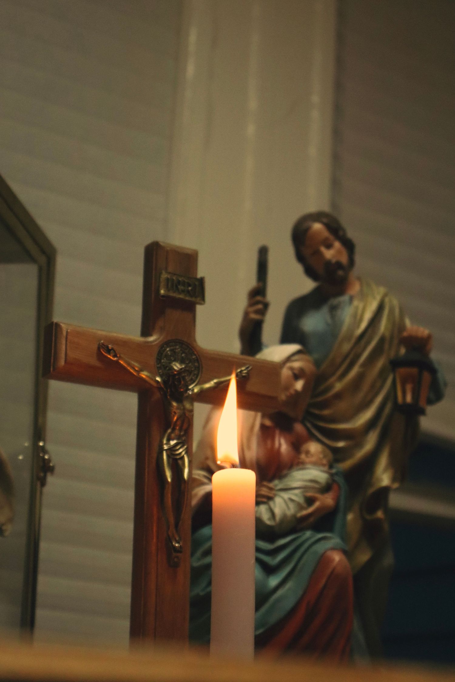 A lit candle glows beside a wooden cross in a dimly lit spiritual or religious setting, creating a peaceful ambiance. Rosary with families