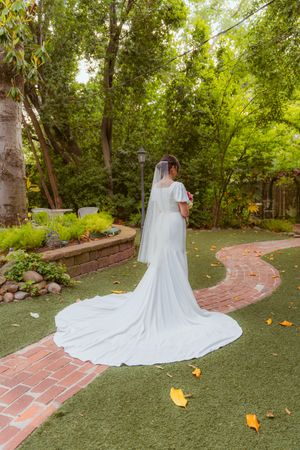 A long white wedding dress with a flowing train on a brick path surrounded by lush garden landscaping.