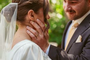 A close-up wedding photo shows hands touching during an intimate moment between newlyweds in an outdoor garden setting.