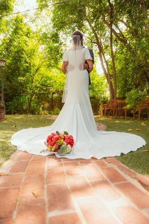 A wedding dress with a flowing train and bouquet of red flowers on a brick patio surrounded by lush greenery.