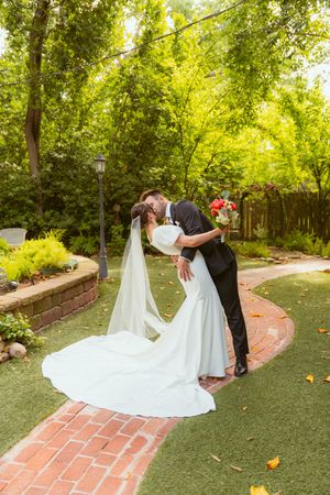 A couple shares a romantic kiss on a brick path surrounded by lush gardens and greenery in a charming outdoor setting.
