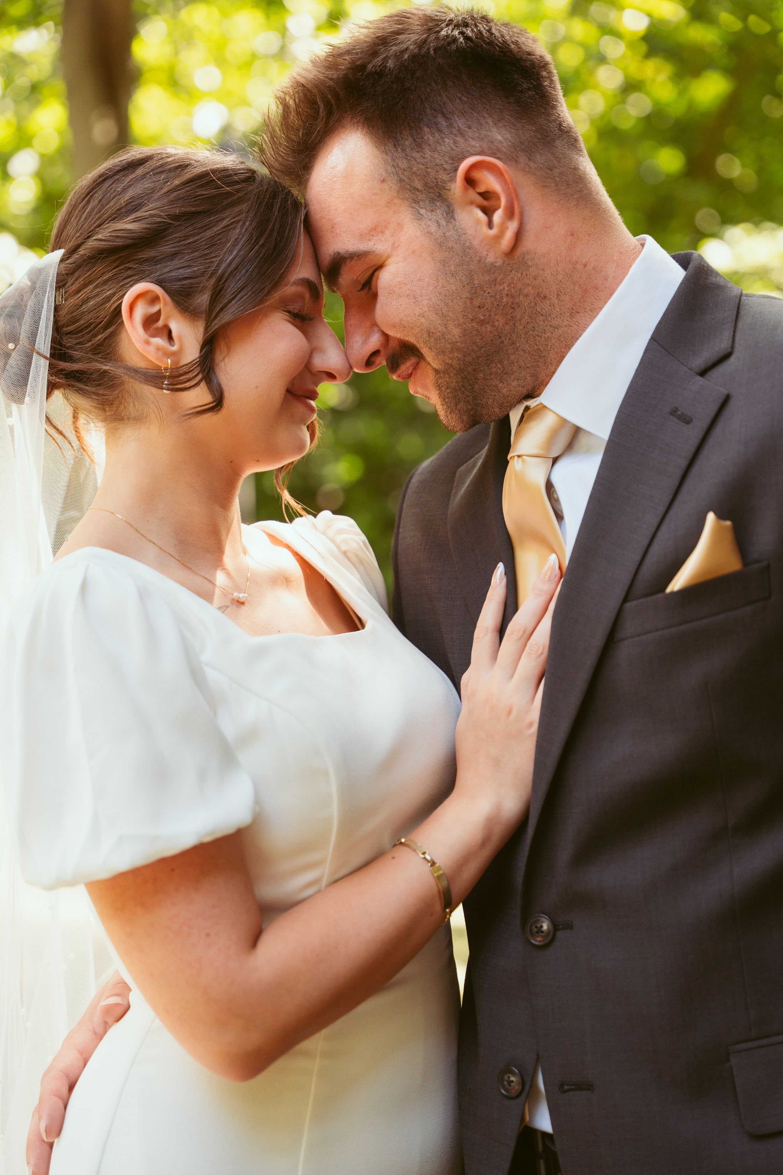 A tender romantic moment between a bride in white and groom in dark suit embracing outdoors with sunlit greenery behind them.