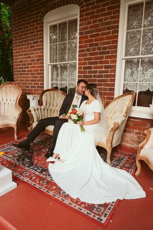 Newlyweds sitting together on vintage chairs on a red brick porch with lace-curtained windows.