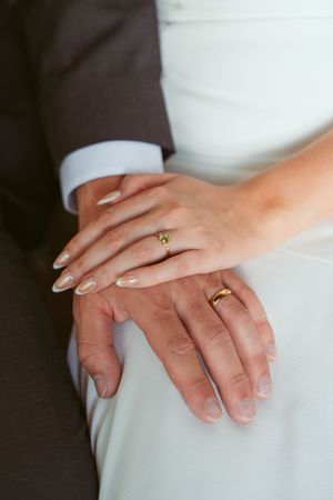 Close-up shot of newlywed couple's hands with wedding rings against white dress and dark suit backdrop.