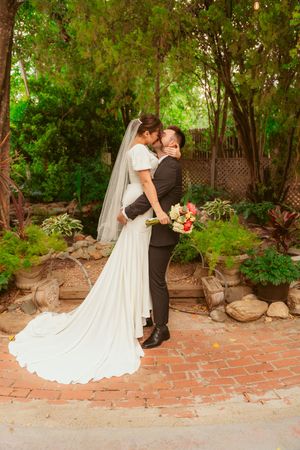 Newlyweds share an intimate moment on brick patio with lush greenery and trailing white wedding gown.