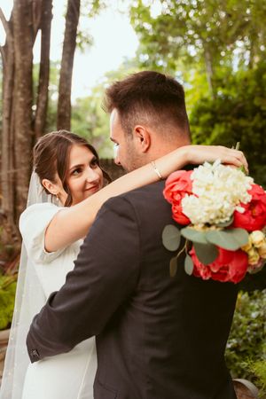 Bride embraces groom from behind while holding vibrant red and white wedding bouquet in outdoor forest setting.