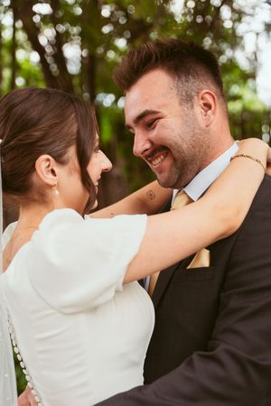 A bride and groom share an intimate moment outdoors with joyful smiles while embracing in their wedding attire.