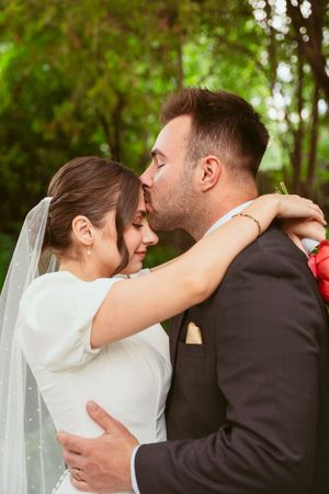 A romantic wedding couple embraces in an outdoor garden setting with the bride wearing a white dress and flowing veil.
