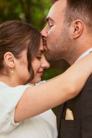 Intimate moment between newlyweds as groom kisses bride's forehead in outdoor garden setting.
