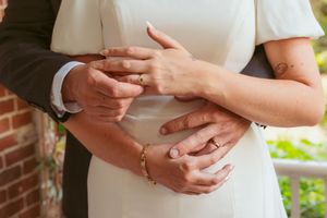 Close-up of hands showing a wrapped engagement ring and wedding bands against white dress and brick wall background.