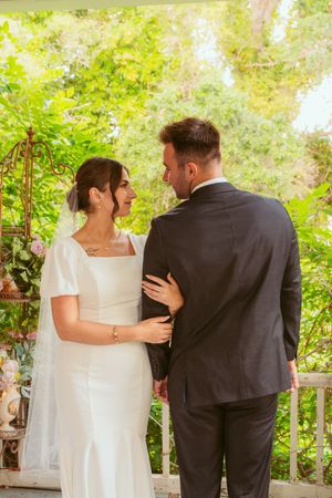 Couple in wedding attire embracing on an outdoor porch surrounded by greenery.