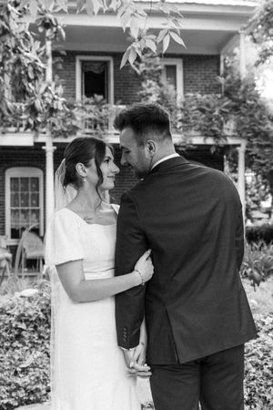Black and white wedding photograph showing couple in elegant attire standing together in front of historic brick house.